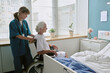 © AnnaStills - Young adult Caucasian female nurse assisting senior Caucasian woman sitting in wheelchair near hospital bed, nurse standing behind patient, medical care setting visible through window