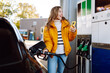 © maxbelchenko - A young woman in a bright jacket fills up her car at a gas station. A beautiful woman with a phone fills up her car via an app. Concept of transport, gas stations and technology.