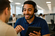 © Jens - Interactive Collaboration: An engaged customer service representative smiles warmly as he interacts with a customer, holding a tablet, with other professionals in the background.