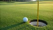 © Phaimanee - A white golf ball on a lush green golf course. The ball is resting on the short grass of the putting green, just inches away from the hole. The hole itself is visible, with a yellow flagstick planted