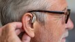 © Enigma - Senior male wearing glasses, carefully positioning digital hearing aid for enhanced auditory communication and personal connection