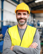 © Kowit - A smiling man in a yellow hard hat and safety vest stands confidently in an industrial setting, ready for work.