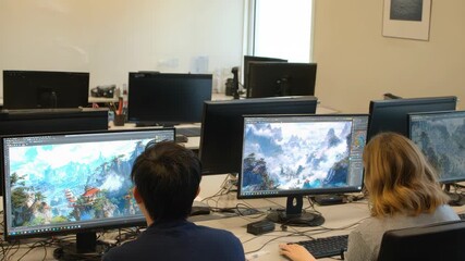 Two individuals work on computers with landscape art displayed on their screens suggesting graphic design game development or digital art