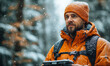 © Bartek - Confident man wearing orange beanie and jacket hiking in snowy forest holding remote control device for outdoor exploration in winter landscape