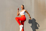 Fit woman exercising outdoors, raising leg and jumping, doing functional training outside on bright sunny day, wearing sport outfit