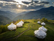 © AmazingAerialAgency - Aerial view of white domed tents perch on a vibrant green hillside, nestled beneath rolling mountains under a sky streaked with dramatic clouds, Tago, Adjara, Georgia.
