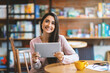 © Prostock-studio - Happy arab woman sitting at cafe, using digital tablet and drinking coffee, smiling at camera, copy space. Young latin lady holding digital pad, blogging while resting at cafeteria, working online