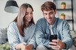 © Daniel - Happy millennial couple sit relax on couch in living room watching video on cellphone together, smiling young husband and wife rest on sofa at home browsing Internet using modern phone device