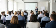 © Renderis - Audience listens to a speaker presenting on a screen in a bright room.