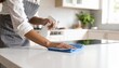 © Rupa - Woman in an apron cleaning a modern white kitchen countertop with a spray bottle and a blue microfiber cloth.