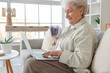 © Pixel-Shot - Senior woman with glass cup of butterfly pea flower tea and laptop sitting on sofa in living room