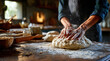 © David Giraud - Mains pétrissant de la pâte à pain sur une table en bois, entourées de farine et d'ustensiles de cuisine, symbolisant le savoir-faire artisanal de la boulangerie traditionnelle, vue de léger profil.