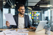 © Liubomir - A smiling businessman reviews documents while working on a laptop in a modern office environment, focused on productivity.