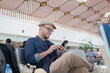 © Reezky - Cheerful young Asian man sitting on a bench while holding smartphone, waiting at airport lounge.