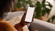 © Johannes - young black african woman holding smartphone in hand with white black empty mockup screen template using cell phone relaxing on sofa at home over shoulder view mobile apps advertising concept no logo