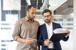 © Liubomir - Two business professionals, one holding a tablet, the other examining a document, engaged in a discussion within a modern office setting.