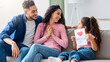 © Prostock-studio - Happy Mother's Day Concept. Smiling little girl holding handmade greeting card with drawn red heart for her mom, congratulating her mommy while they sitting together on couch in living room