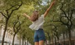© DhanaStudio - Young woman with outstretched arms enjoying urban park with green trees