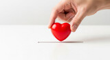 A person's hand gently placing a small red heart on a white surface, perfect for illustrating concepts of giving, care, or a small act of kindness