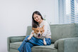 © NanSan - A sweet Asian woman relaxes at home, gently petting her happy corgi on the sofa. Their cozy bonding time reflects warmth, friendship, love for animals, and peaceful indoor moments filled with smiles