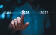 © Brishy  - Businessman's hand pointing at a glowing virtual timeline, starting the new year 2026. Concept of future planning, business goals and vision.