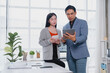 © Tj - Two asian businesspeople are discussing and working together using tablet and calculator in the brightly lit office with plants and supplies on the desk