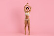 © Anastasiya - Confident young black woman posing in beige underwear, smiling with arms raised. Studio shot on pink background shows her toned and fit body in natural light, full length