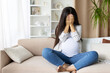 © Anastasiya - Pregnant woman sitting cross legged on couch, covering face with hands, crying at home expressing anxiety, fear, or overwhelming emotion. Honest portrayal of pregnancy mood swings