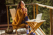 © vita - Relaxing woman on a wooden balcony enjoying coffee and working on laptop in the forest setting. A calm girl enjoys tea drink and sunlight on a wooden terrace, wrapped in a cozy blanket enjoying warmth