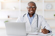 © Prostock-studio - Friendly african american doctor working with laptop in modern office, taking notes, planning his day. Man smiling to camera, sitting at workplace in office