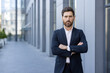 © Liubomir - An assertive businessman wearing a suit confidently poses outdoors in an urban setting. The image conveys professionalism, confidence, and leadership in a contemporary business environment.
