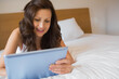 © WavebreakMediaMicro - Woman in late twenties lying on bedroom bed with white linens using tablet leaning on pillow