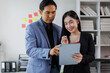 © David - Two asian coworkers standing in the office discuss about business plan and looking at tablet.handsome asia man and beautiful asian woman as business partners working at indoor