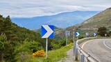 Panoramic view of the road in the mountains. Travelling by car.Winding mountain road with metal guardrails and blue arrow traffic signs. Lush green vegetation lines the roadside, with yellow flowering