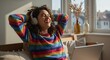 © rdkcho - Joyful Young Woman Relaxing at Home, Smiling with Headphones On and Arms Behind Her Head in a Cozy Living Space with Window Light