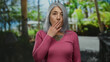 © Krakenimages.com - Senior woman with grey hair in a park setting outdoors expressing surprise or thoughtfulness, wearing a pink sweater against a lush green backdrop with trees and plants visible.
