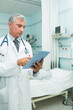 © wavebreak3 - Senior male doctor standing in ward wearing lab coat and using tablet, copy space