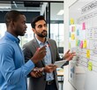 © picturehome - Two diverse professional men, project managers working on task standing next to large white board with attached sticky notes, analyzing schedule, discussing strategy, engaged in productive teamwork