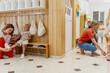 © lordn - Mother helping her son change shoes in a preschool locker area while the teacher assists another child nearby.
