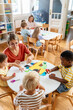 © lordn - Preschool teacher interacting with a diverse group of children during an arts and crafts activity, fostering creativity and multicultural learning.