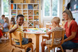 © lordn - Smiling boy waving at the camera while sitting with classmates and teacher during a multicultural preschool craft activity.