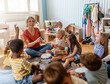 © lordn - Preschool teacher leading a fun interactive activity with children sitting in a circle, raising hands and smiling in a bright classroom