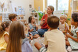 © lordn - Preschool teacher leading a fun interactive activity with children sitting in a circle, raising hands and smiling in a bright classroom