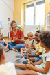 © lordn - Preschool teacher leading a fun interactive activity with children sitting in a circle, raising hands and smiling in a bright classroom
