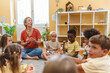 © lordn - Preschool teacher leading a fun interactive activity with children sitting in a circle, raising hands and smiling in a bright classroom
