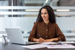 © Liubomir - A professional woman sits at a desk, smiling while working on papers and using her laptop.