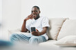 © SHOTPRIME STUDIO - Happy young African American man laughing while using a smartphone at home, wearing casual white t shirt and jeans in a bright, minimalistic living room