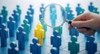 © bryanwisnu - A hand holding a magnifying glass inspects a single blue figure in a crowd, symbolizing recruitment, search, and human resources.
