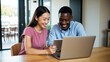 © MT Studio - A smiling asian woman and african american man looking at a laptop and tablet on a wooden table