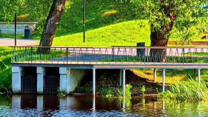 Wall Mural - wooden pedestrian bridge over calm river in green park with trees and grass
